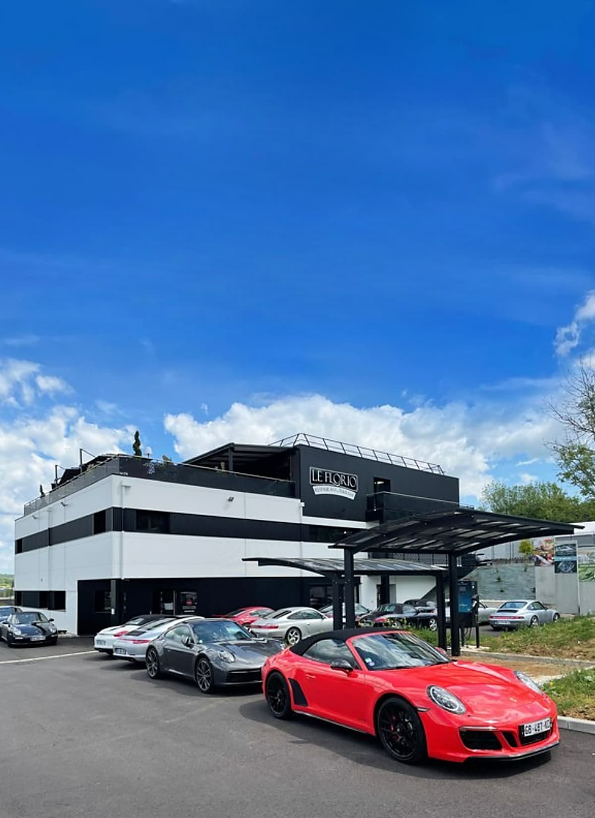 Façade du showroom sous un ciel bleu, avec une Porsche 911 Carrera GTS cabriolet rouge au premier plan